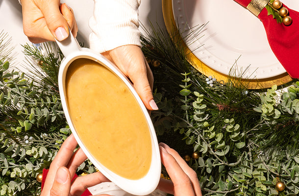 Someone passing a gravy boat of Kevin's Natural Foods Turkey Gravy over the top of an arrangement of Christmas decorations.